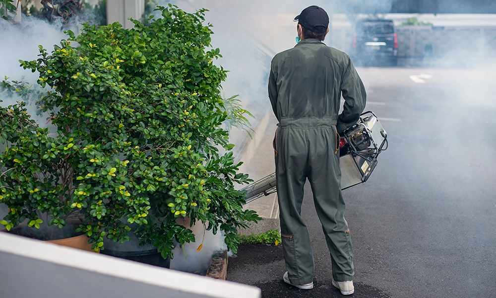 A pest control worker in a protective suit using a fogging machine to spray chemicals for mosquito control outdoors, surrounded by greenery and smoke.