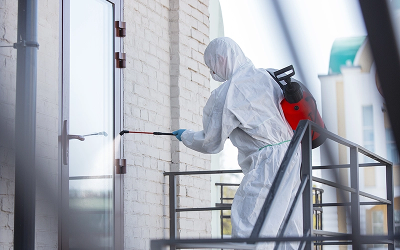 A pest control professional wearing protective gear sprays insecticide on the exterior of a building to manage and prevent pest infestations.