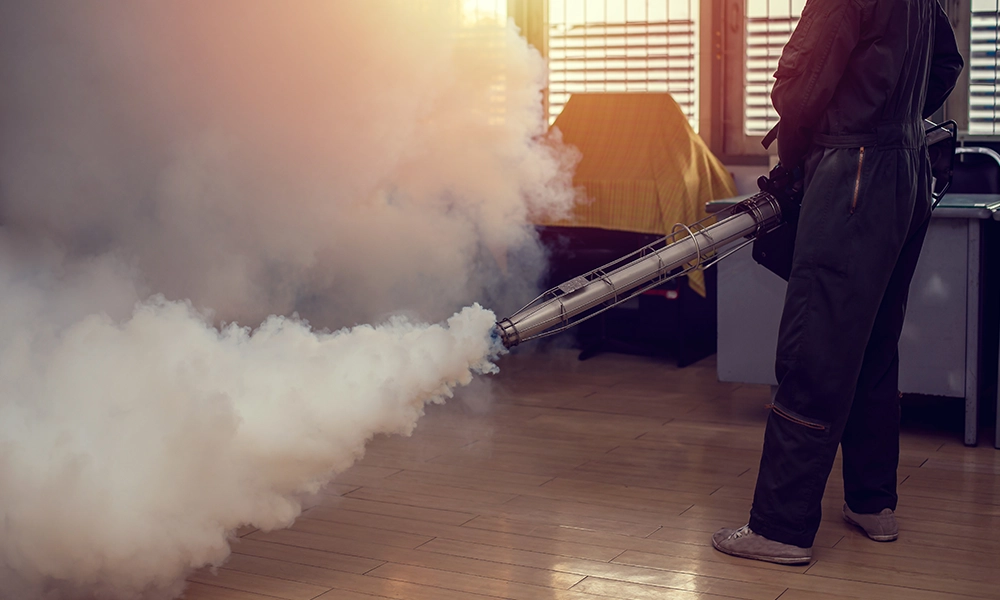 A pest control technician using a fogging machine to release thick clouds of smoke indoors, as part of a mosquito control treatment, with sunlight streaming through the windows in the background.