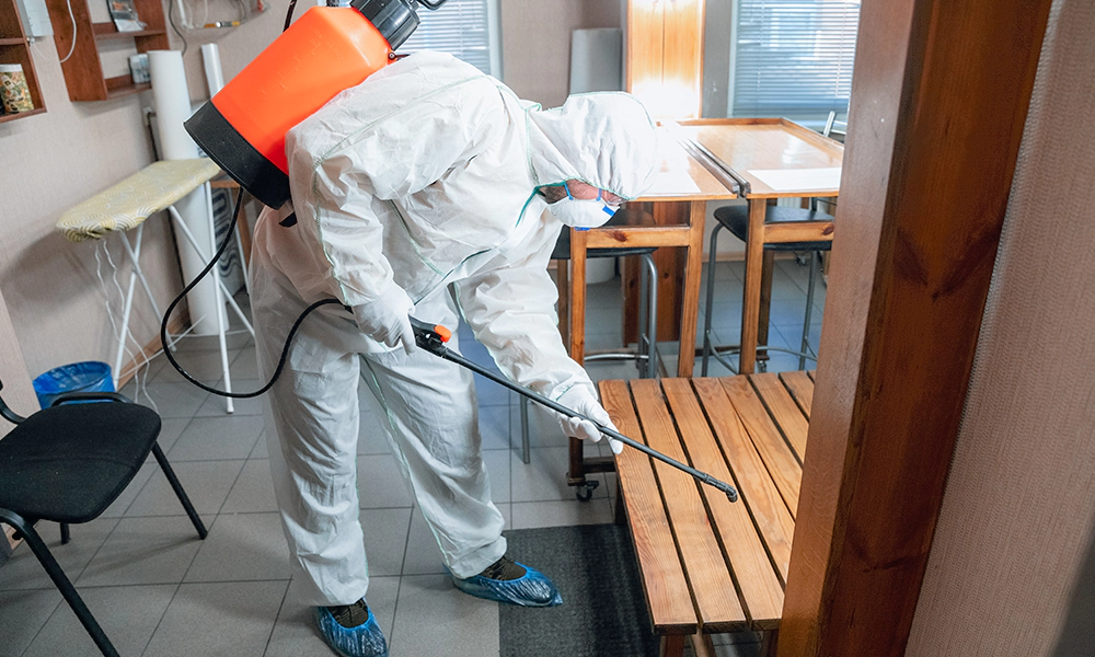 A pest control professional wearing protective gear uses a sprayer to apply treatment inside a residential home to prevent pests.