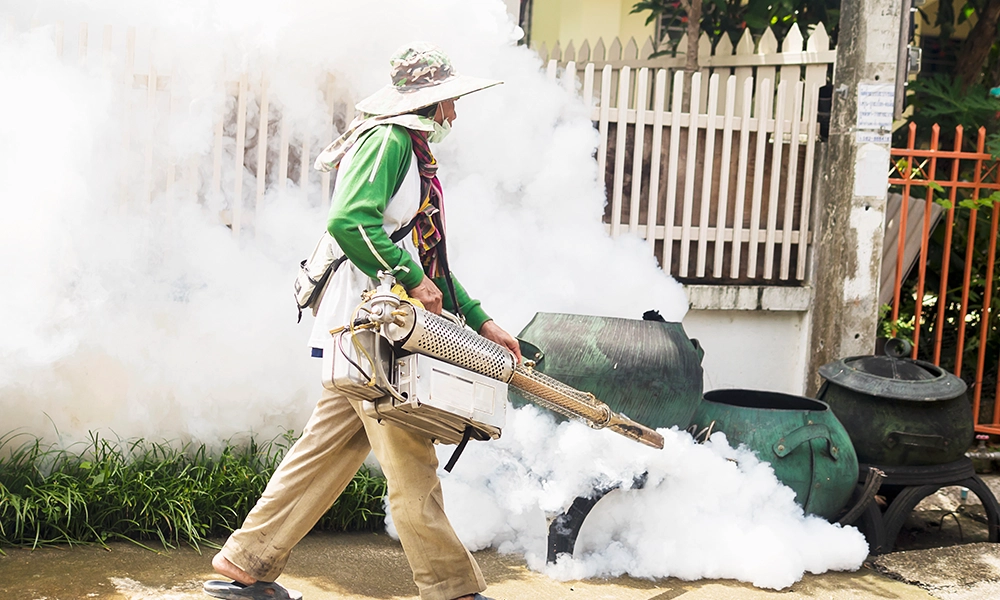 A pest control worker using a fogging machine for mosquito control outside a property, surrounded by thick white smoke.