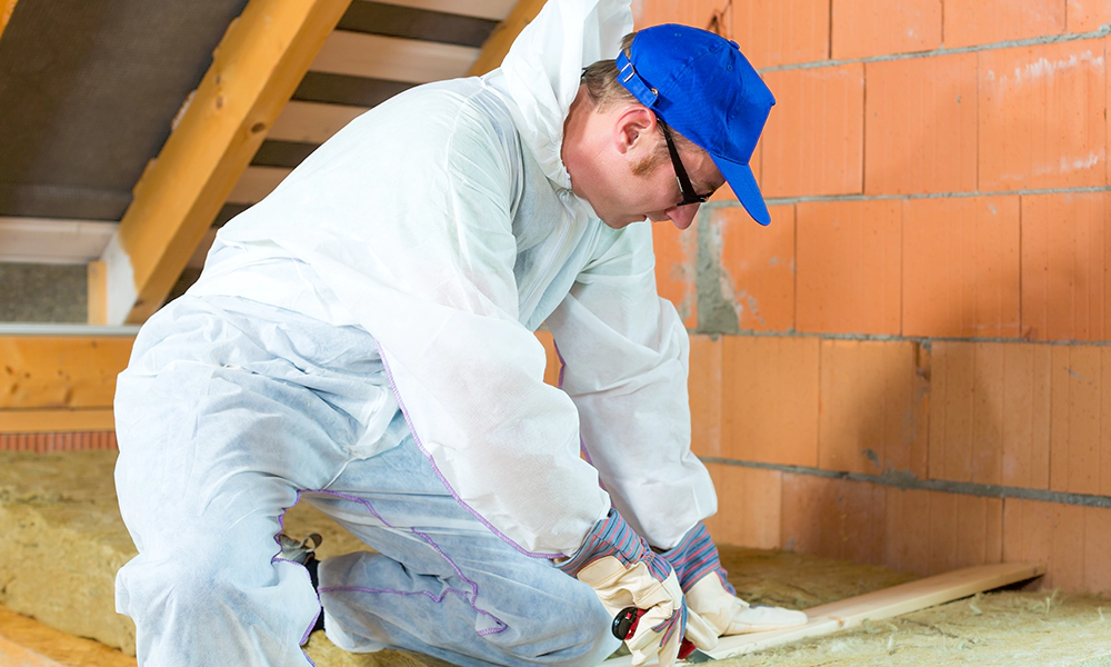 A worker wearing protective gear cuts insulation material in an attic, demonstrating home insulation installation.