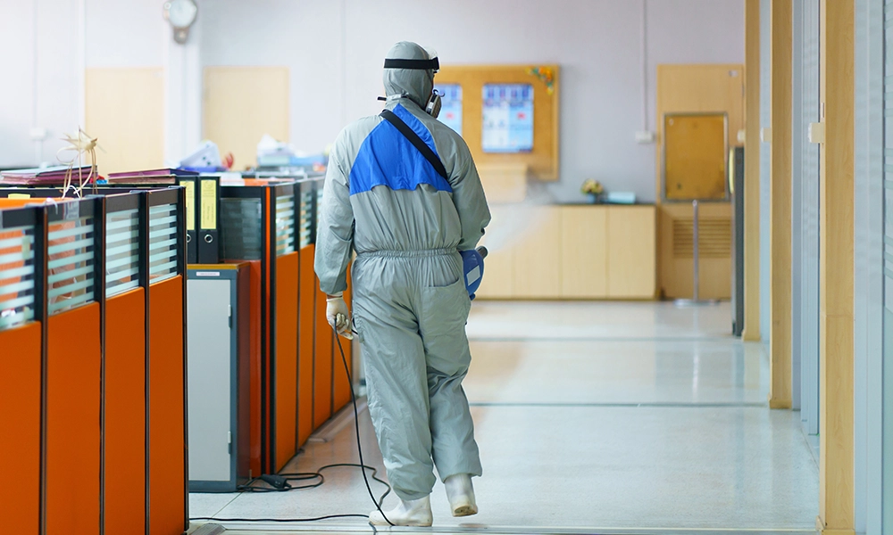 A pest control specialist in protective gear sprays an office hallway, taking preventive measures against pests in a workplace setting.