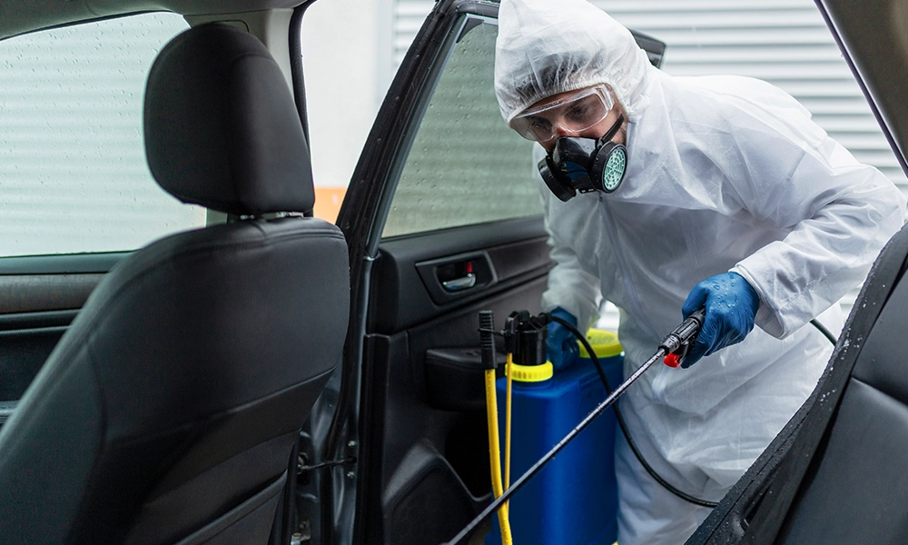 A professional in full protective gear, including a mask and gloves, uses a sprayer to disinfect and treat the interior of a car, focusing on bed bug removal and sanitation.