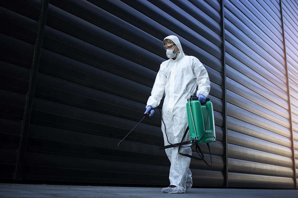 A pest control professional in full protective gear and mask, holding a green spray container and walking alongside a large industrial building, performing pest control measures to prevent infestations.
