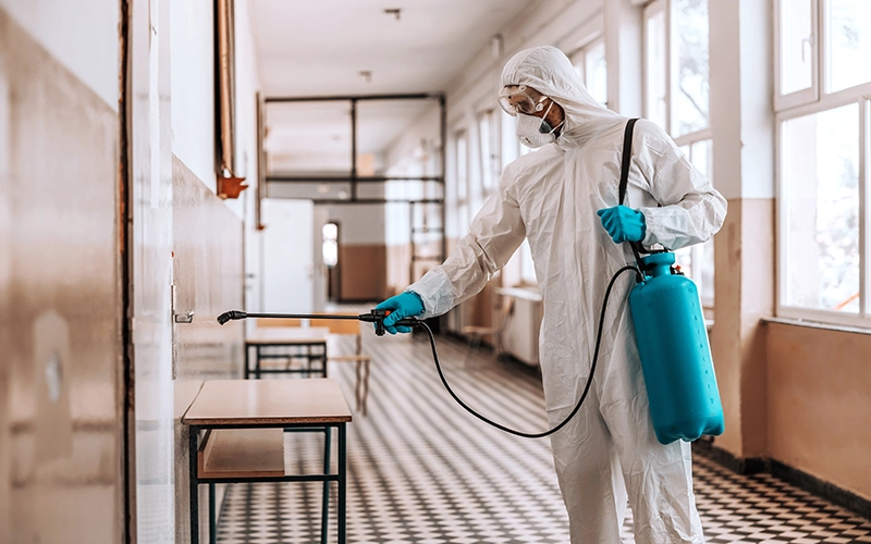A pest control professional in full protective suit sprays a classroom area, ensuring a safe and pest-free learning environment.