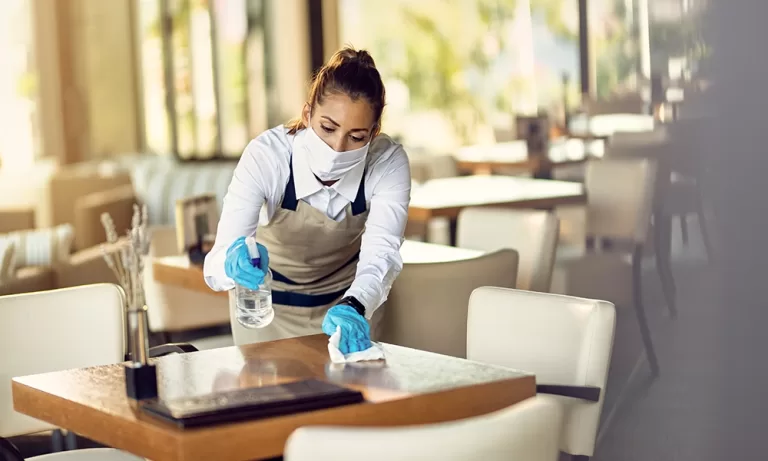 A restaurant worker wearing a face mask and gloves diligently sanitizes a table using a spray bottle and cloth, emphasizing hygiene and cleanliness. This image highlights the importance of implementing an Integrated Pest Management (IPM) program for maintaining a safe and sanitary restaurant environment.