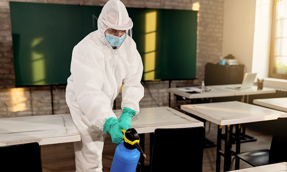 Person wearing full protective gear, including a white hazmat suit, face mask, and gloves, spraying a disinfectant in an empty classroom to prevent or eliminate pests, with desks and a green chalkboard in the background.