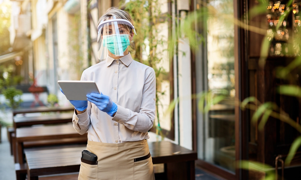 A restaurant worker wearing a face shield, mask, gloves, and an apron, holding a tablet, symbolizing health and safety measures in food service establishments.